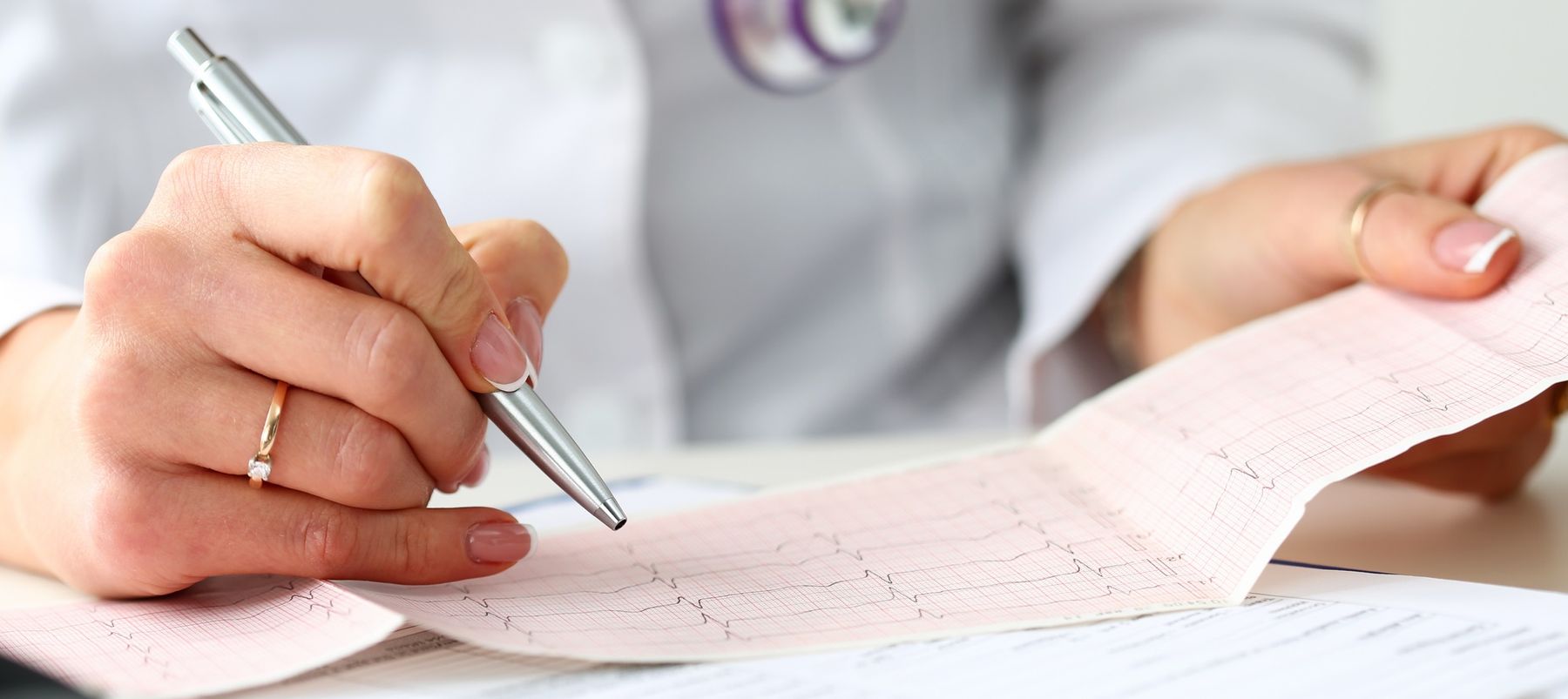 A white coat-wearing doctor holds the results of an EKG test in one hand and traces over them with a pen in the other hand