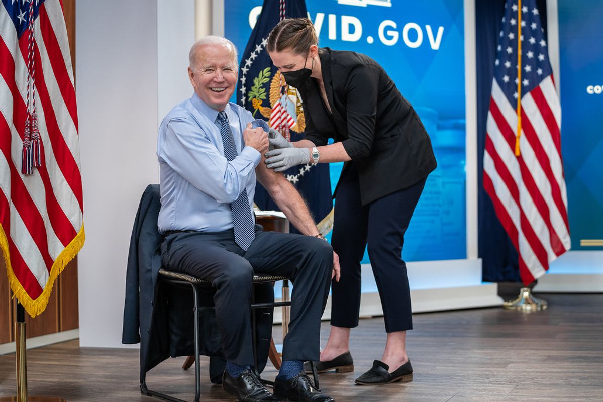 Joe Biden rolling up his sleeve as a healthcare worker leans over him to inject him with a COVID-19 booster shot.