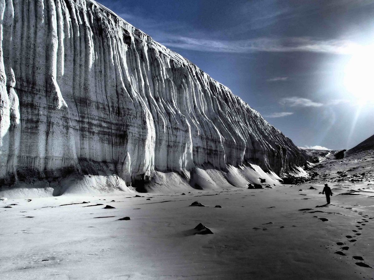 Photo of a distant figure walking away from the camera through a snowy plain. The towering wall of a glacier looms overhead.