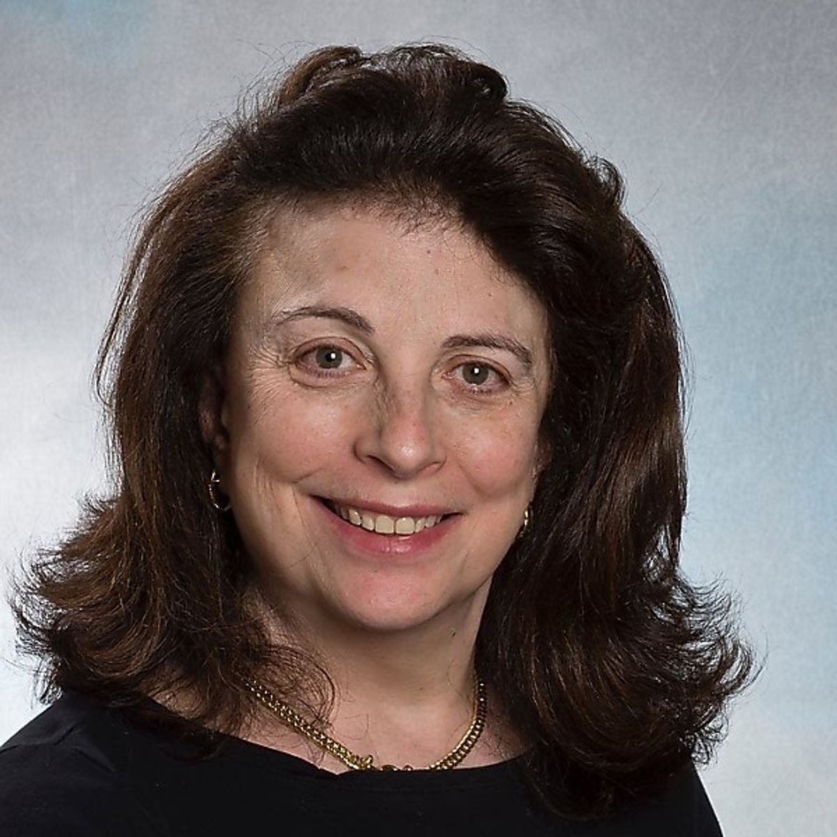 A portrait shot of Beryl Benacerraf, who wears a black shirt and gold necklace and smiles into the camera, on a bluish gray background.