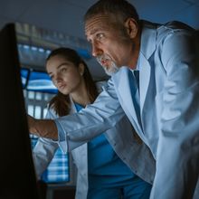 a middle-aged male scientist wearing a white lab coat points at a computer screen while a younger woman scientist also wearing a lab coat looks on.