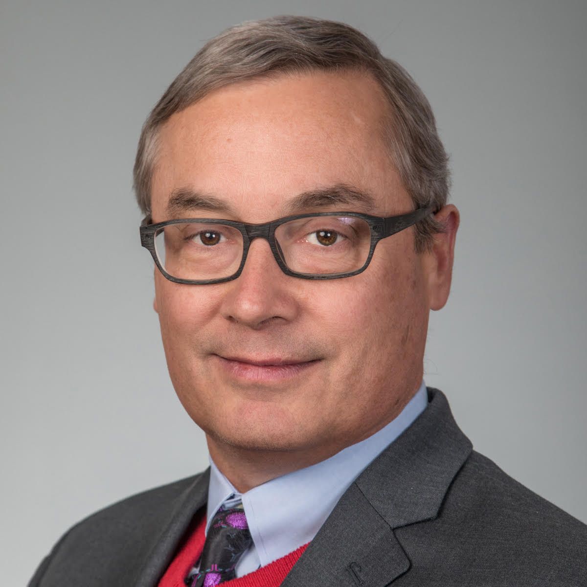 A picture of man with light skin and grey hair, wearing a grey suit jacket, a light blue shirt, a piece of red knitwear and a purple and grey tie.