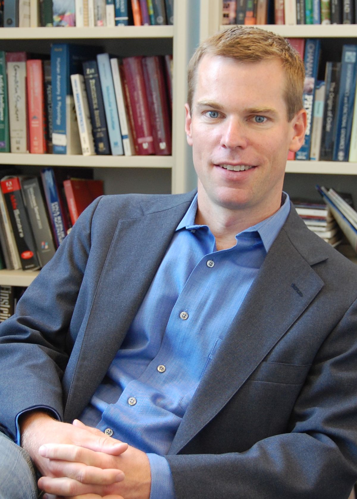 A man wearing grey suit sits in front of a bookshelf. 