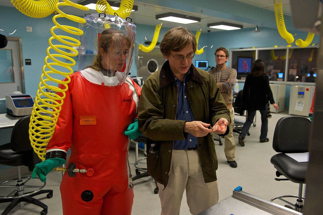 A photograph of Ian Lipkin (right) in a green jacket instructing Jennifer Ehle (left) in a red BSL-4 costume on a scene on the set of Contagion. The set resembles a BSL-4, with yellow, coiled airlines hanging across the ceiling and one connected to Ehle’s suit. A photograph of Ian Lipkin (right) in a green jacket instructing Jennifer Ehle (left) in a red BSL-4 costume on a scene on the set of Contagion. The set resembles a BSL-4, with yellow, coiled airlines hanging across the ceiling and one connected to Ehle’s suit.