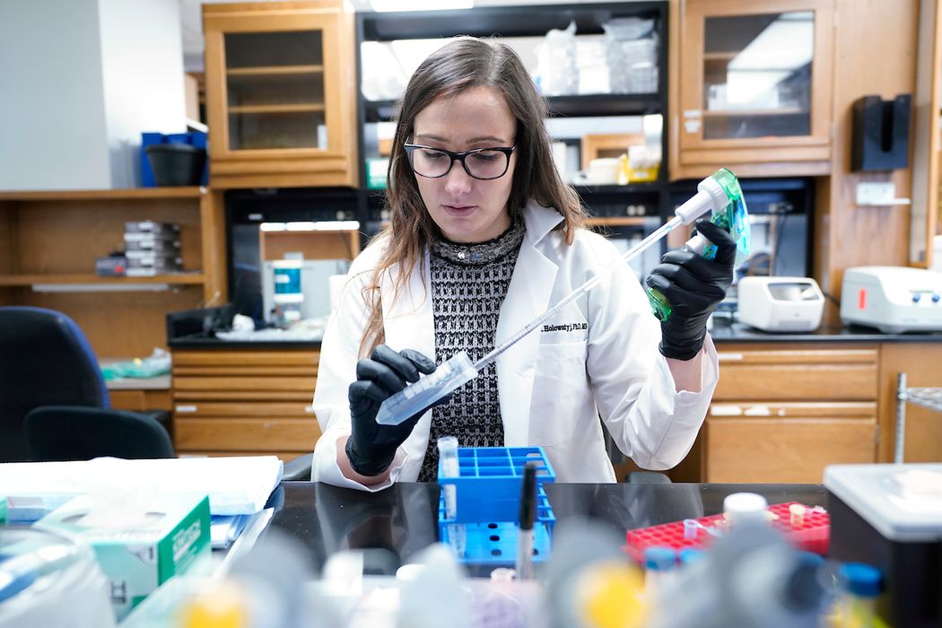 Photo of Holowatyj wearing a white lab coat. She is sitting at a laboratory bench and pipetting a liquid from a falcon tube.