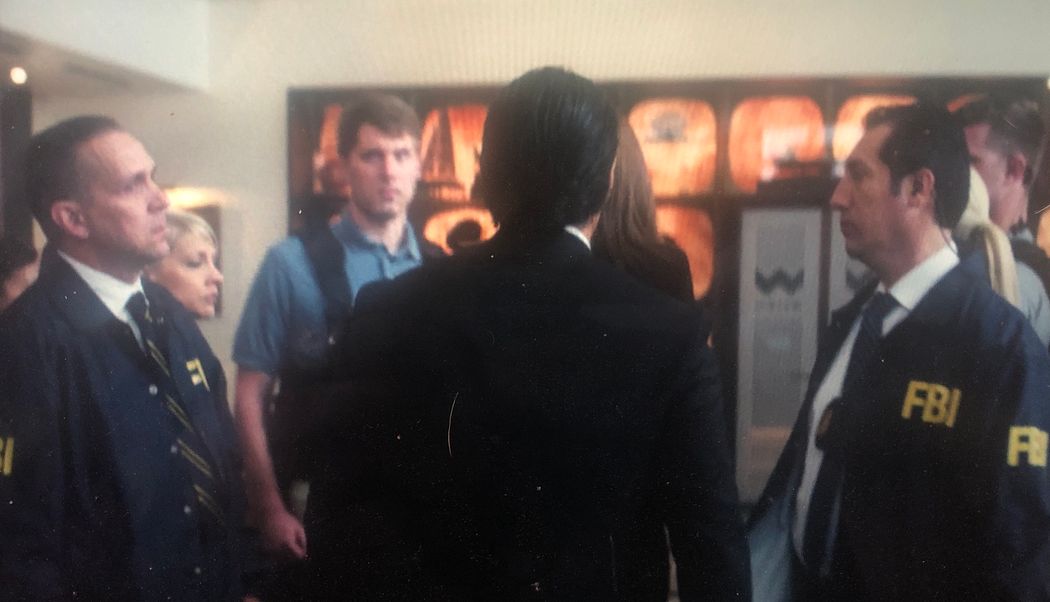 An image of a frame from the movie Rampage showing James Dahlman, a biomedical engineer, playing an FBI agent. Dahlman, unfocused in the background, is wearing a blue shirt and black tactical vest. The focus is on the back of another character’s head, walking toward the group of agents. An image of a frame from the movie Rampage showing James Dahlman, a biomedical engineer, playing an FBI agent. Dahlman, unfocused in the background, is wearing a blue shirt and black tactical vest. The focus is on the back of another character’s head, walking toward the group of agents.