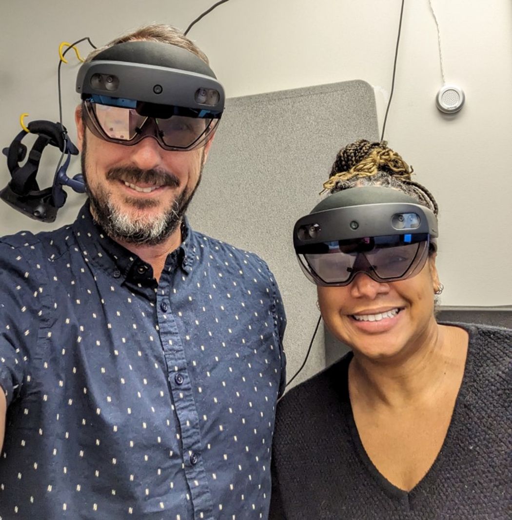 Travis Baker (left, wearing a blue shirt) and Jaleesa Stringfellow (right, wearing a black shirt) smile at the camera while wearing AR headsets. Travis Baker (left, wearing a blue shirt) and Jaleesa Stringfellow (right, wearing a black shirt) smile at the camera while wearing AR headsets.
