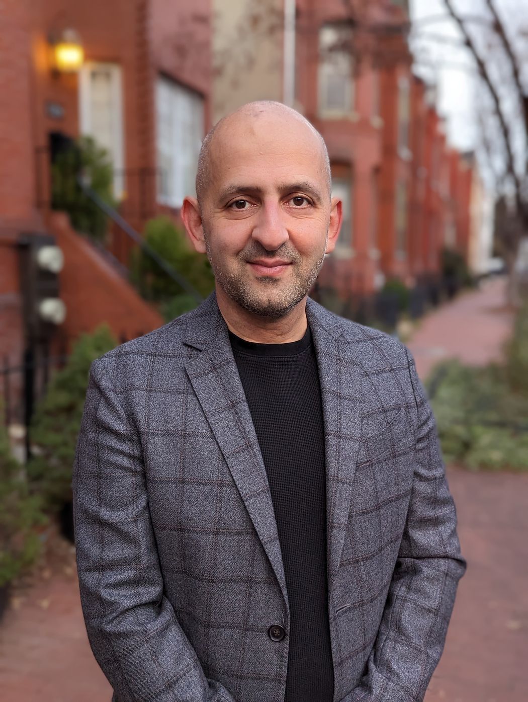 Photograph of Arash Javanbakht standing on the sidewalk by red brick townhomes. He is bald and is wearing a dark shirt and gray dress jacket. Photograph of Arash Javanbakht standing on the sidewalk by red brick townhomes. He is bald and is wearing a dark shirt and gray dress jacket.