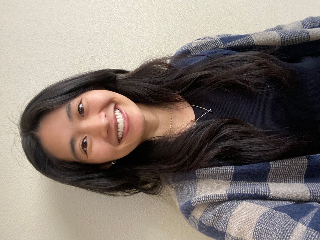 Photograph of Laura Tran. She is smiling at the camera and has long dark hair. She is wearing a dark blue shirt and a blue plaid cardigan.