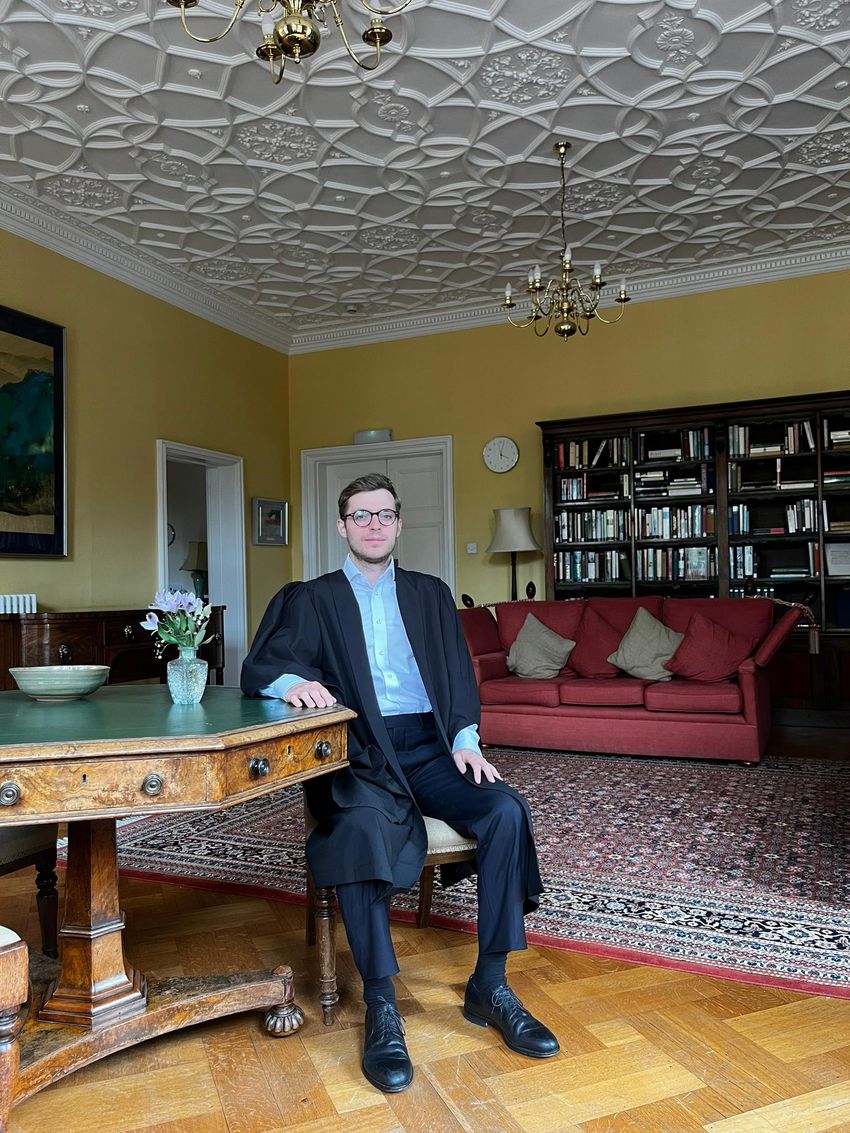 Photograph of Leonardo Bonetti sitting at a wooden table in a room with yellow walls. Bonetti is wearing navy slacks, a long navy dress coat, and a light blue collared shirt. Photograph of Leonardo Bonetti sitting at a wooden table in a room with yellow walls. Bonetti is wearing navy slacks, a long navy dress coat, and a light blue collared shirt.