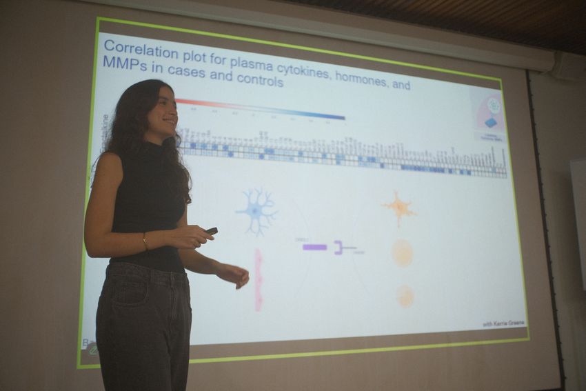 Photograph of Victoria Bastos, a physician scientist and former graduate student at the Federal University of Rio de Janeiro, presenting her work exploring subtypes of ME/CFS. She stands on the left side of the image, wearing dark pants and a dark sleeveless top, in front of a projector showing a presentation slide. She has tan skin and dark hair and is smiling to someone off frame. Photograph of Victoria Bastos, a physician scientist and former graduate student at the Federal University of Rio de Janeiro, presenting her work exploring subtypes of ME/CFS. She stands on the left side of the image, wearing dark pants and a dark sleeveless top, in front of a projector showing a presentation slide. She has tan skin and dark hair and is smiling to someone off frame.