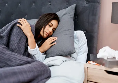 Photo of a woman with straight dark hair looking fatigued lying in bed with a grey blanket covering her. 