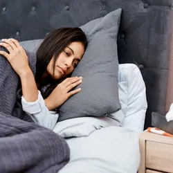 Photo of a woman with straight dark hair looking fatigued lying in bed with a grey blanket covering her. Photo of a woman with straight dark hair looking fatigued lying in bed with a grey blanket covering her.