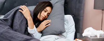 Photo of a woman with straight dark hair looking fatigued lying in bed with a grey blanket covering her. 
