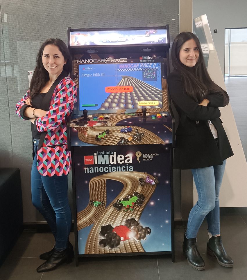 Photograph of Patricia Bondia (left) in a red patterned blazer and jeans and Melek Villanueva (right) in a black blazer and jeans standing on either side of an arcade game featuring their video game, Nanocar Racing: The Game. Photograph of Patricia Bondia (left) in a red patterned blazer and jeans and Melek Villanueva (right) in a black blazer and jeans standing on either side of an arcade game featuring their video game, Nanocar Racing: The Game.
