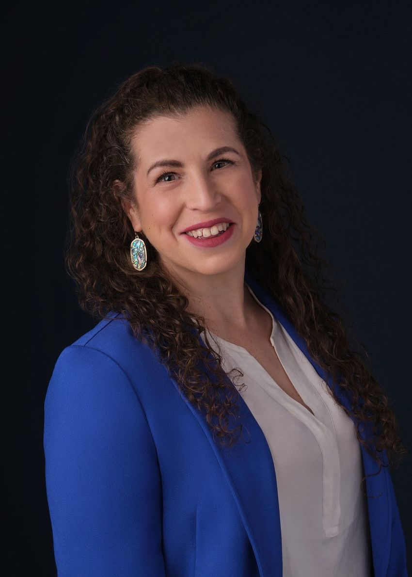 A smiling woman with dark, curly brown hair wearing a white blouse and a royal blue blazer. A smiling woman with dark, curly brown hair wearing a white blouse and a royal blue blazer.