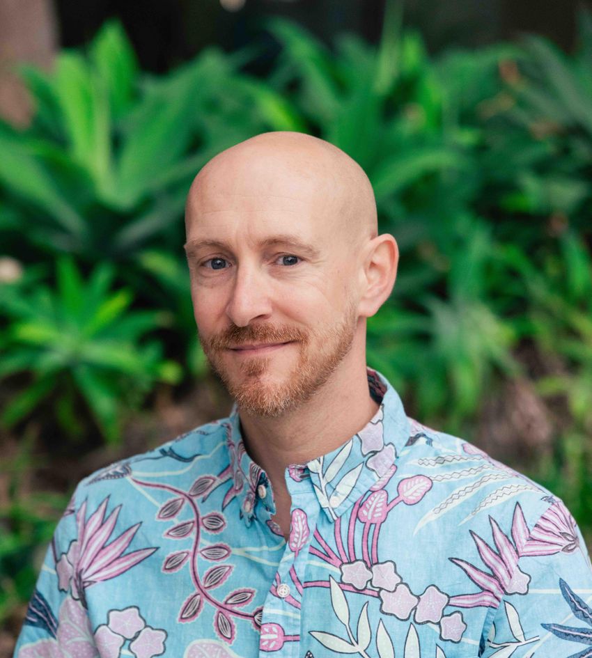Image of Philip Gordts, wearing a light blue shirt with pink botanical patterns, standing in front of a blurred background with plants. Image of Philip Gordts, wearing a light blue shirt with pink botanical patterns, standing in front of a blurred background with plants.