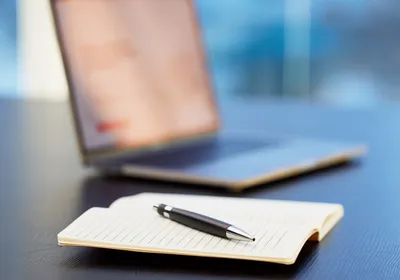 Closeup image of an open notebook with a pen in the foreground and an open laptop computer in the background.