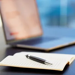 Closeup image of an open notebook with a pen in the foreground and an open laptop computer in the background.