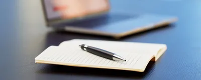 Closeup image of an open notebook with a pen in the foreground and an open laptop computer in the background.