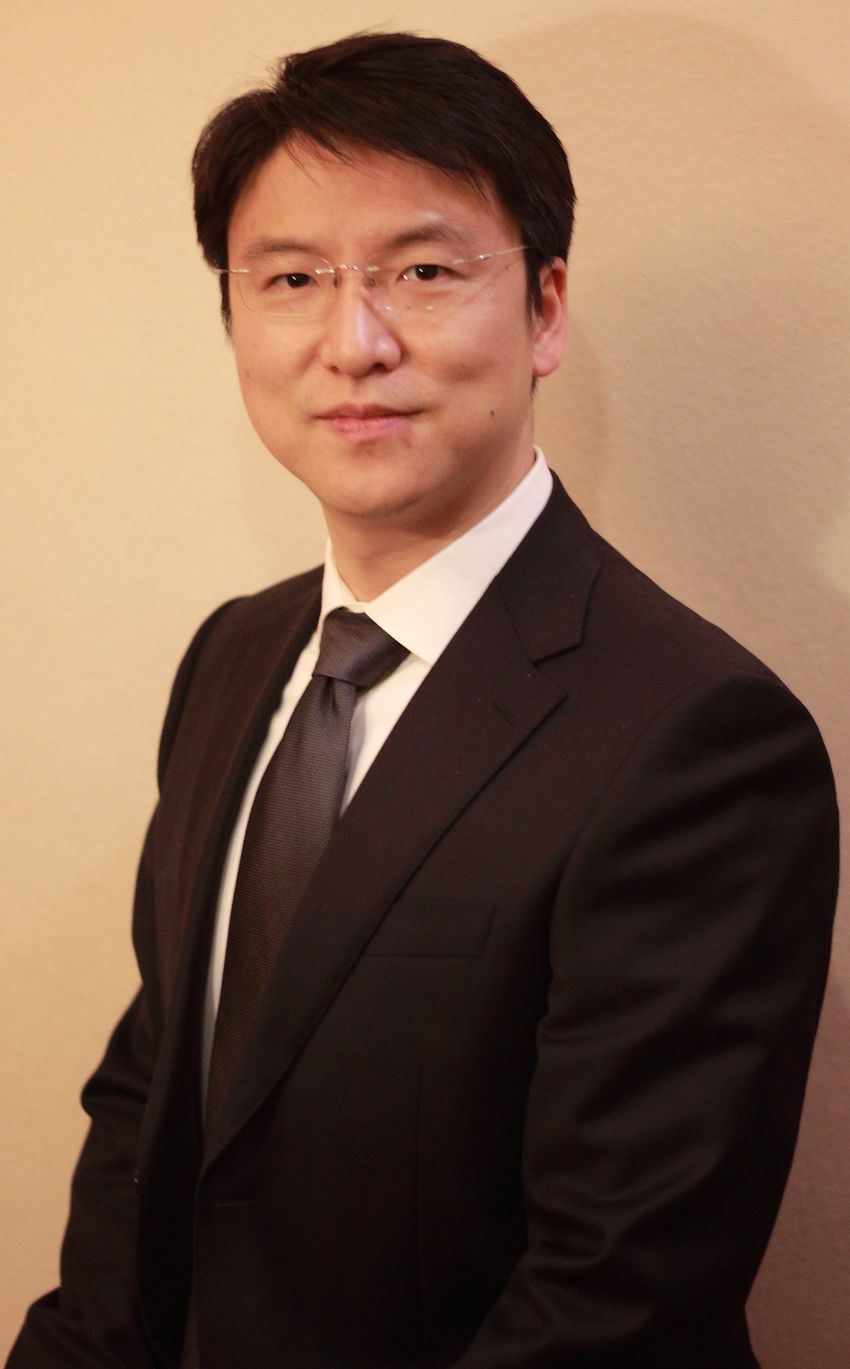 Image of Xing Chen, wearing glasses and a black suit, on a light yellow background Image of Xing Chen, wearing glasses and a black suit, on a light yellow background