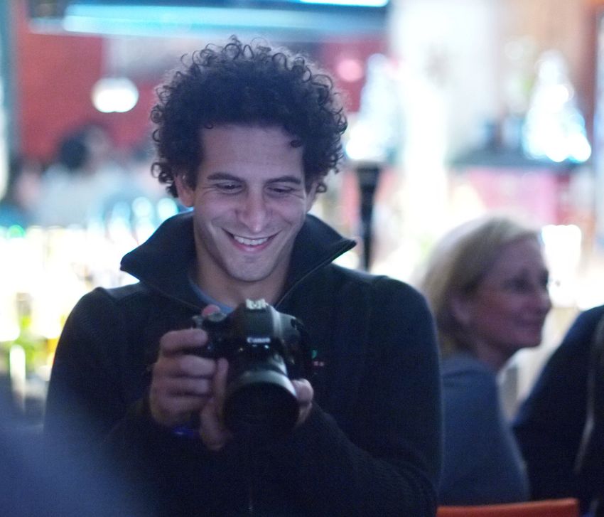 A man with long curly brown hair smiles and looks down at a camera.