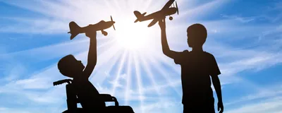 The silhouettes of two children, one standing and one in a wheelchair, playing with toy aeroplanes against a sunny blue sky.