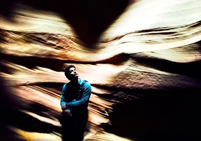A man with dark hair wears a blue shirt with his arms crossed as he leans against the wall of a slot canyon.