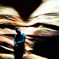 A man with dark hair wears a blue shirt with his arms crossed as he leans against the wall of a slot canyon.