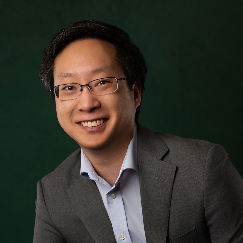 A photo of Brian Lin wearing a grey blazer and blue shirt against a green background. He wears glasses and smiles at the camera.