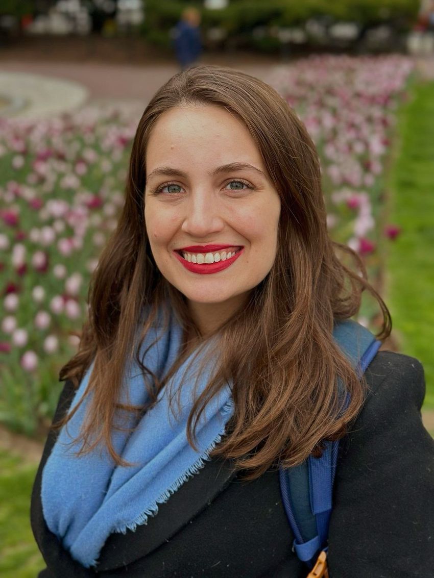 A photo of Juliana Gutschow Gameiro in front of flowers. She wears a black coat and blue scarf and smiles at the camera.