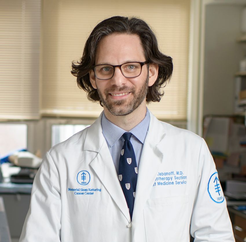 Image of Christopher Klebanoff in front of the blurred background of a room. He’s wearing glasses, a light blue shirt with a dark blue patterned tie, and a buttoned white lab coat embroidered with his name and his insitution’s name and logo. Image of Christopher Klebanoff in front of the blurred background of a room. He’s wearing glasses, a light blue shirt with a dark blue patterned tie, and a buttoned white lab coat embroidered with his name and his insitution’s name and logo.
