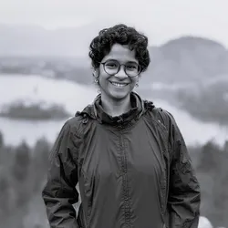 Photograph of Sahana Sitaraman. The photograph is in grayscale. Sahana has short, curly hair, round-framed glasses, and is wearing a windbreaker jacket. Photograph of Sahana Sitaraman. The photograph is in grayscale. Sahana has short, curly hair, round-framed glasses, and is wearing a windbreaker jacket.