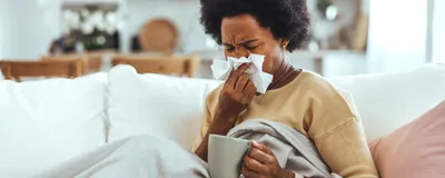 A young, sick woman sitting on a sofa, blowing her nose with a tissue.
