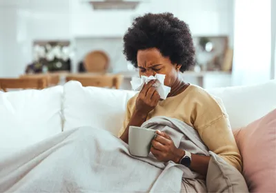 A young, sick woman sitting on a sofa, blowing her nose with a tissue. A young, sick woman sitting on a sofa, blowing her nose with a tissue.