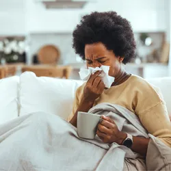 A young, sick woman sitting on a sofa, blowing her nose with a tissue. 