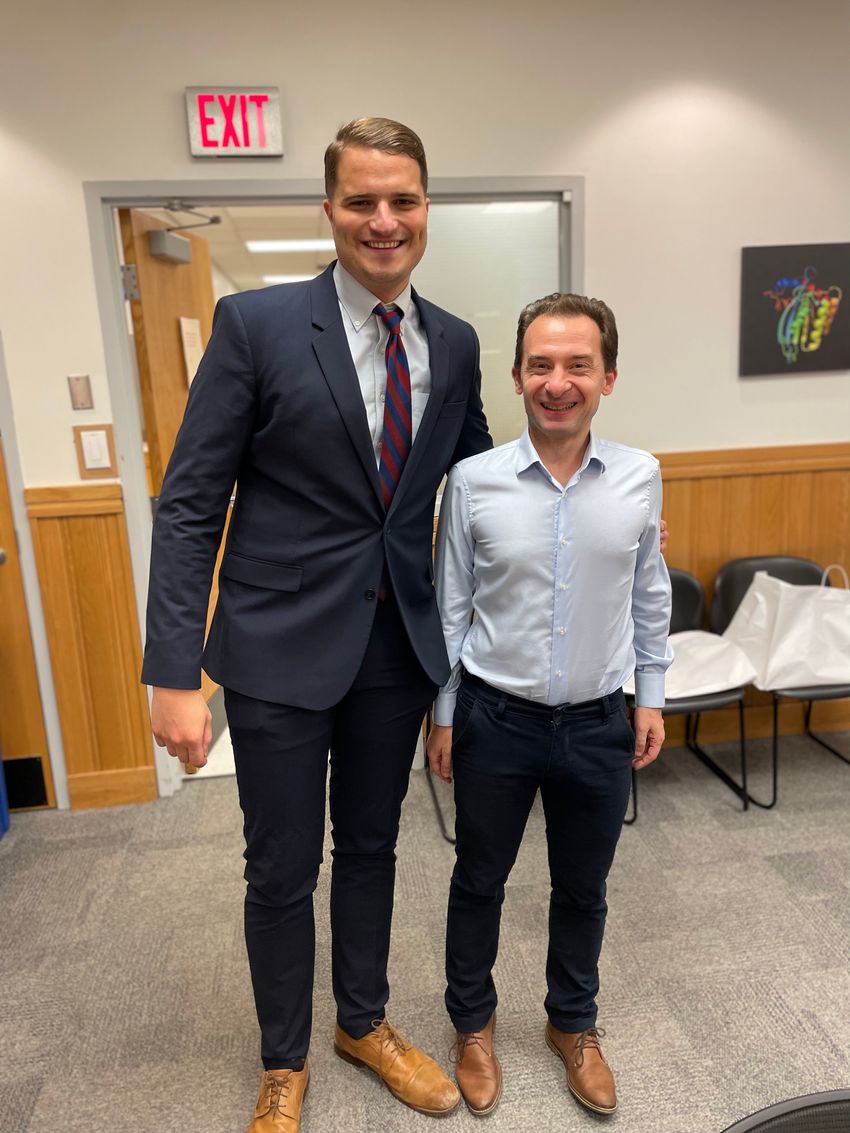Matthew Pine and Drew Weismann standing in front of a wood-paneled walls and an open door designated with an exit sign. Weismann is wearing a light blue shirt, dark pants, and brown shoes. Pine is wearing a navy suit, light blue shirt, red and blue striped tie, and brown shoes and has one arm around Weismann. Matthew Pine and Drew Weismann standing in front of a wood-paneled walls and an open door designated with an exit sign. Weismann is wearing a light blue shirt, dark pants, and brown shoes. Pine is wearing a navy suit, light blue shirt, red and blue striped tie, and brown shoes and has one arm around Weismann.