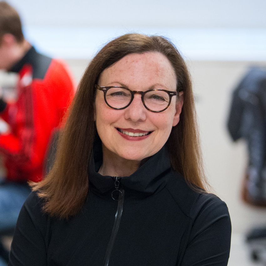 Image of Shelley Berger, wearing glasses and a black zip-up jacket, on a blurred background with a seated individual. Image of Shelley Berger, wearing glasses and a black zip-up jacket, on a blurred background with a seated individual.
