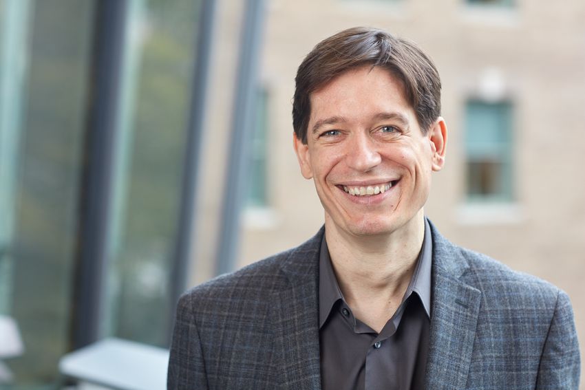 Image of Daniel Kronauer, wearing a dark patterned suit over a grey shirt, on a blurred background of a building. Image of Daniel Kronauer, wearing a dark patterned suit over a grey shirt, on a blurred background of a building.