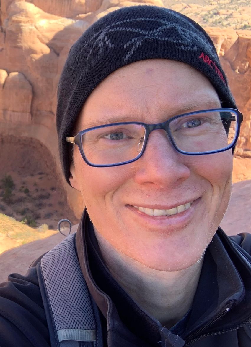Close-up selfie image of Christopher Gregg, wearing glasses, dark patterned hat, a jacket, and a backpack in front of a geological formation. Close-up selfie image of Christopher Gregg, wearing glasses, dark patterned hat, a jacket, and a backpack in front of a geological formation.