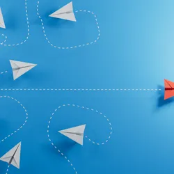 3D illustration of six paper airplanes flying in swirls, depicted by white dashed lines, on the left, and one red airplane flying straight across to the right on a blue background, used here as an indicator for leadership in academia. 