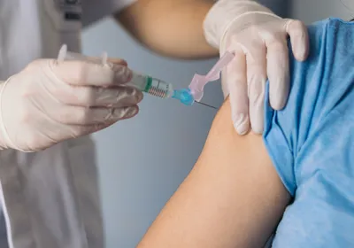 A medical professional, wearing light grey scrubs and white laboratory gloves, immunizes a patient, wearing a blue T-shirt, on their right upper arm.