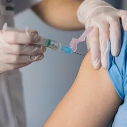 A medical professional, wearing light grey scrubs and white laboratory gloves, immunizes a patient, wearing a blue T-shirt, on their right upper arm.
