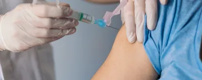 A medical professional, wearing light grey scrubs and white laboratory gloves, immunizes a patient, wearing a blue T-shirt, on their right upper arm.