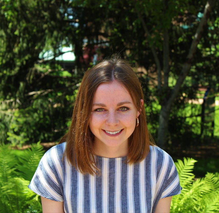 Photo of Katherine Donald, a microbiologist who studies IgA-microbiota interactions, against trees. She wears a blue and white top and smiles at the camera.