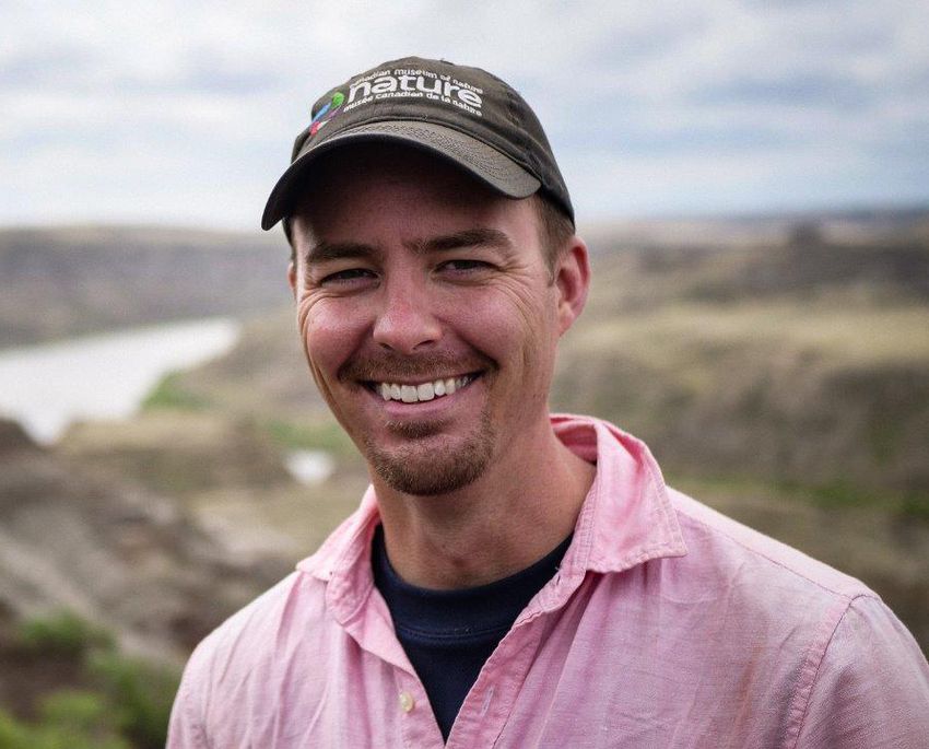 Image of Jordan Mallon, wearing a pink shirt and dark logo cap, on a blurred landscape background.