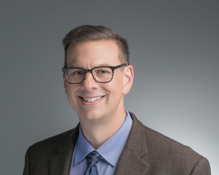 Matthew Girgenti, wearing glasses, a blue shirt, a patterned dark blue tie, and a dark brown patterned suit, poses in front of a grey background.