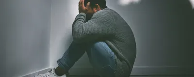 A child with short hair, wearing a grey textured sweater, blue jeans, and white shoes, sits facing the corner of a room. He buries his face in his hands.
