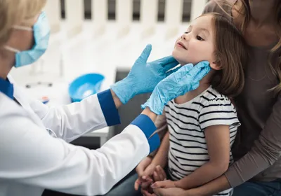 A doctor examines tonsils of a smiling little girl sitting in a woman’s lap.