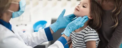 A doctor examines tonsils of a smiling little girl sitting in a woman’s lap.